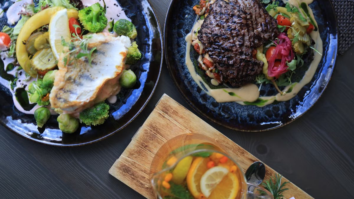 Top view of gourmet fish and steak dishes with fresh citrus and herbs on a stylish table setting.
